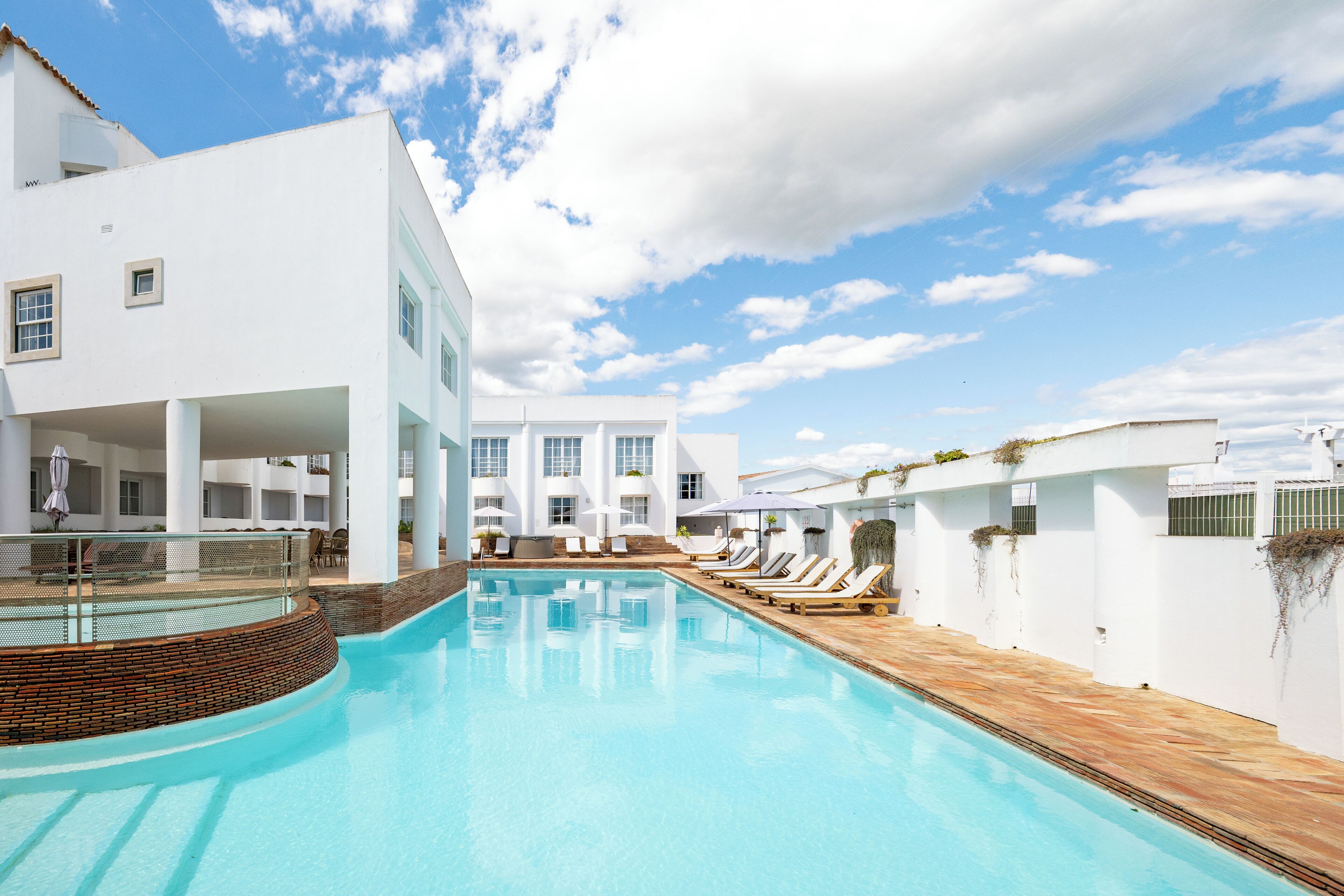 A modern hotel with a swimming pool, sun loungers, and a white exterior.
