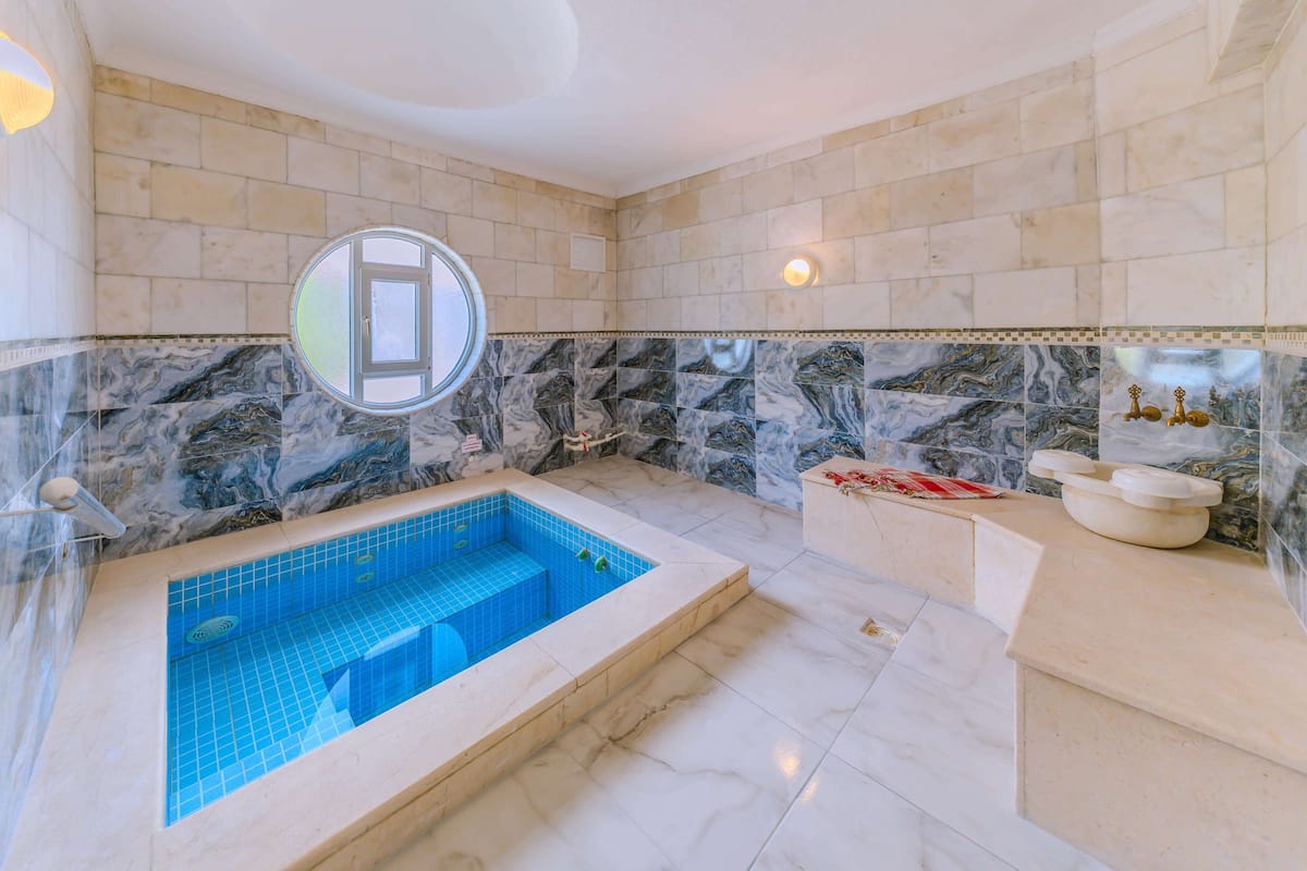 Un baño con pared de mármol, bañera con azulejos azules y espejo redondo.