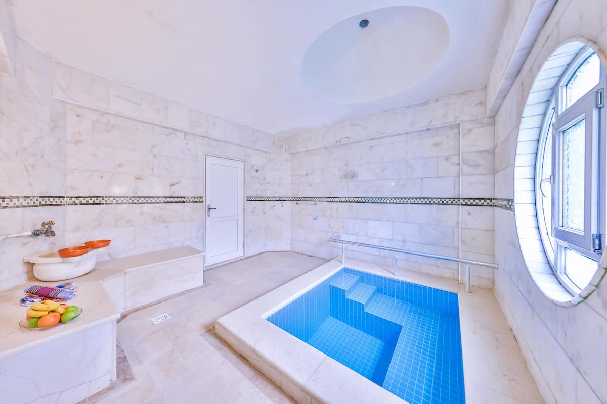 Un baño con piso de mármol, piscina con azulejos azules, lavamanos blanco y una ventana.
