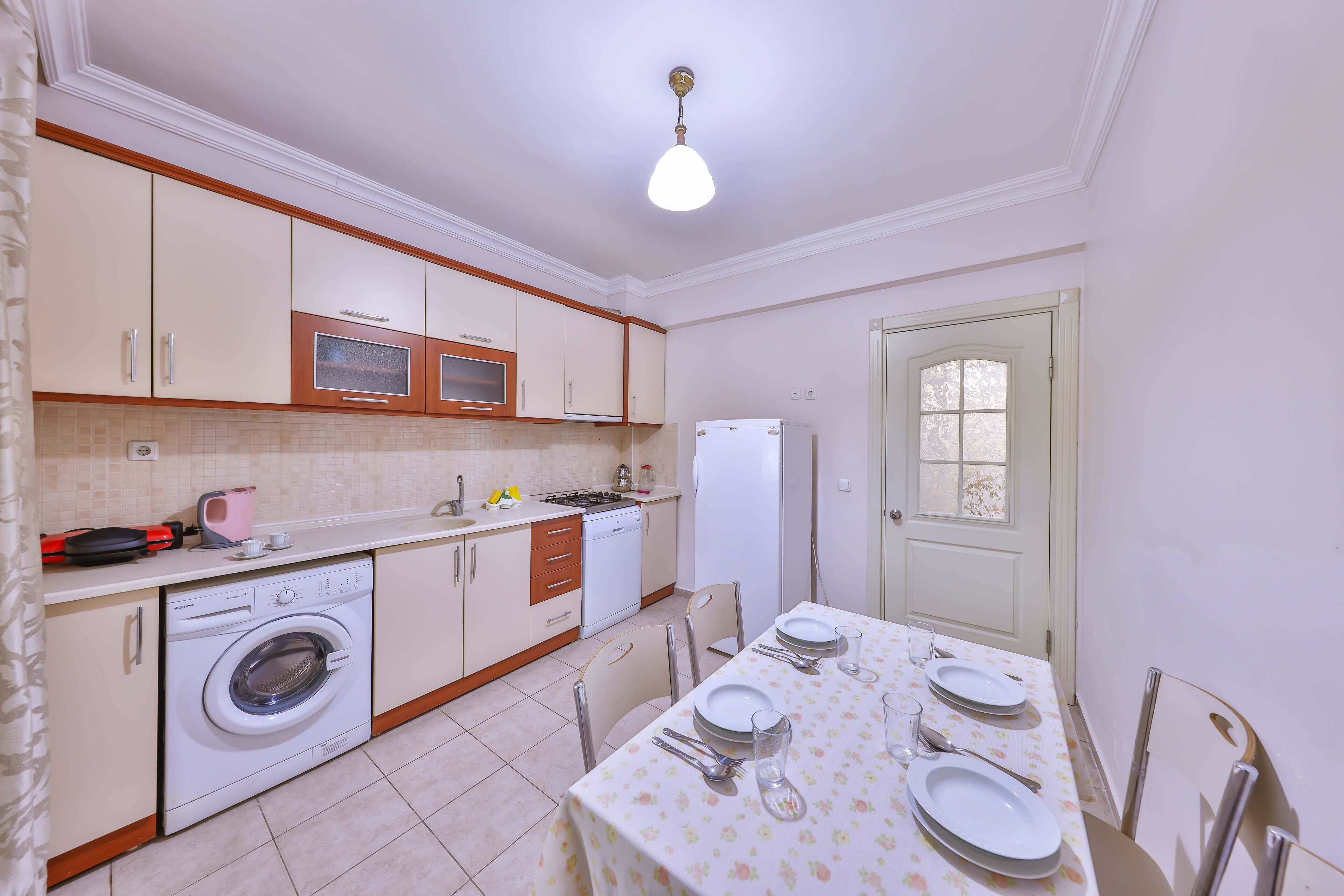 A compact kitchen with a washing machine, microwave, and refrigerator. There is a dining table set with plates and cutlery.