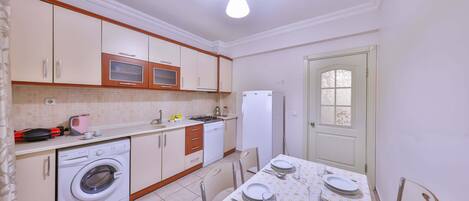 A compact kitchen with a washing machine, microwave, and refrigerator. There is a dining table set with plates and cutlery.