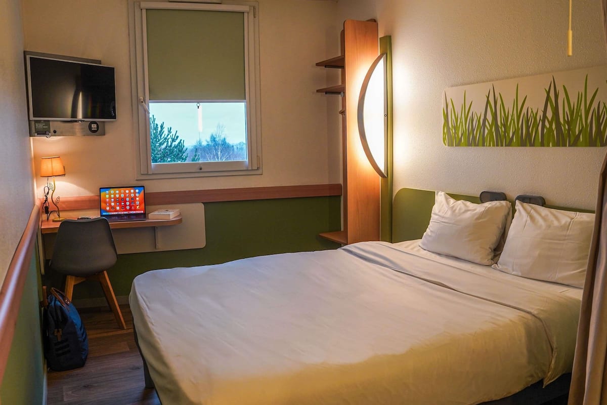A hotel room with a bed, a desk with a laptop, a TV, and a window with a view of trees.