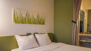 A hotel room with a bed, two pillows, a nightstand, a wall-mounted artwork, and a bathroom visible through a curtain. - ibis budget Bayeux (Nonant)