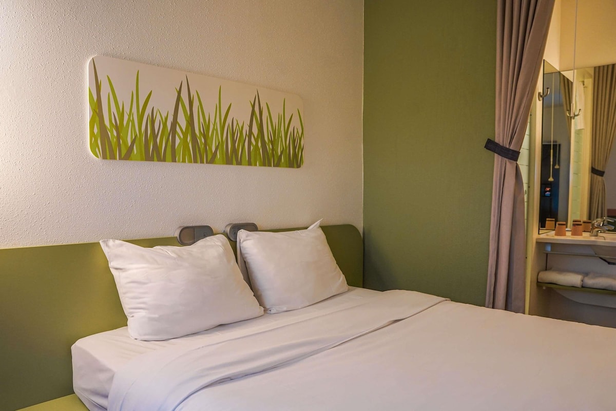 Quarto de hotel com cama, duas almofadas, mesa de cabeceira, obra de arte na parede e banheiro visível por uma cortina.