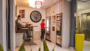 A modern interior with a large wall clock, a vending machine, and a seating area with a person reading a newspaper. - ibis budget Bayeux (Nonant)