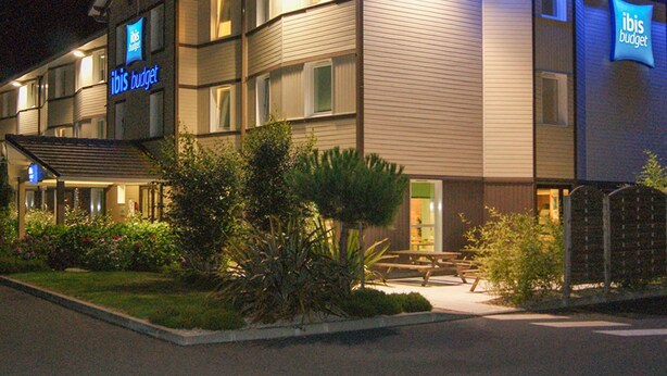 A hotel with a sign reading "ibis budget." It has multiple floors, a covered entrance, and a well-maintained garden area with benches. - ibis budget Bayeux (Nonant)