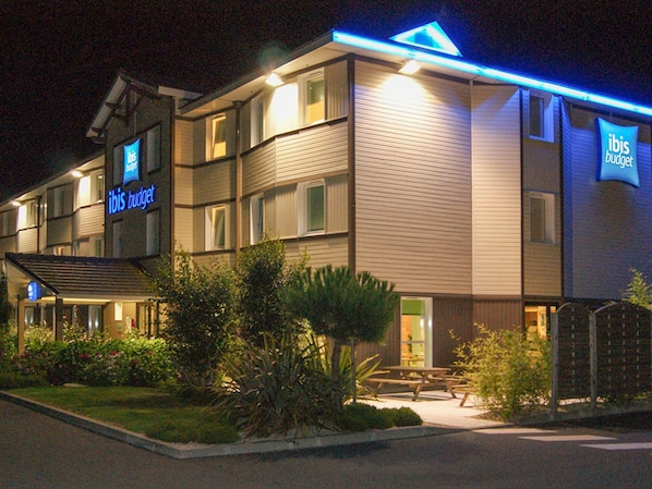 A hotel with a sign reading "ibis budget." It has multiple floors, a covered entrance, and a well-maintained garden area with benches. - ibis budget Bayeux (Nonant)