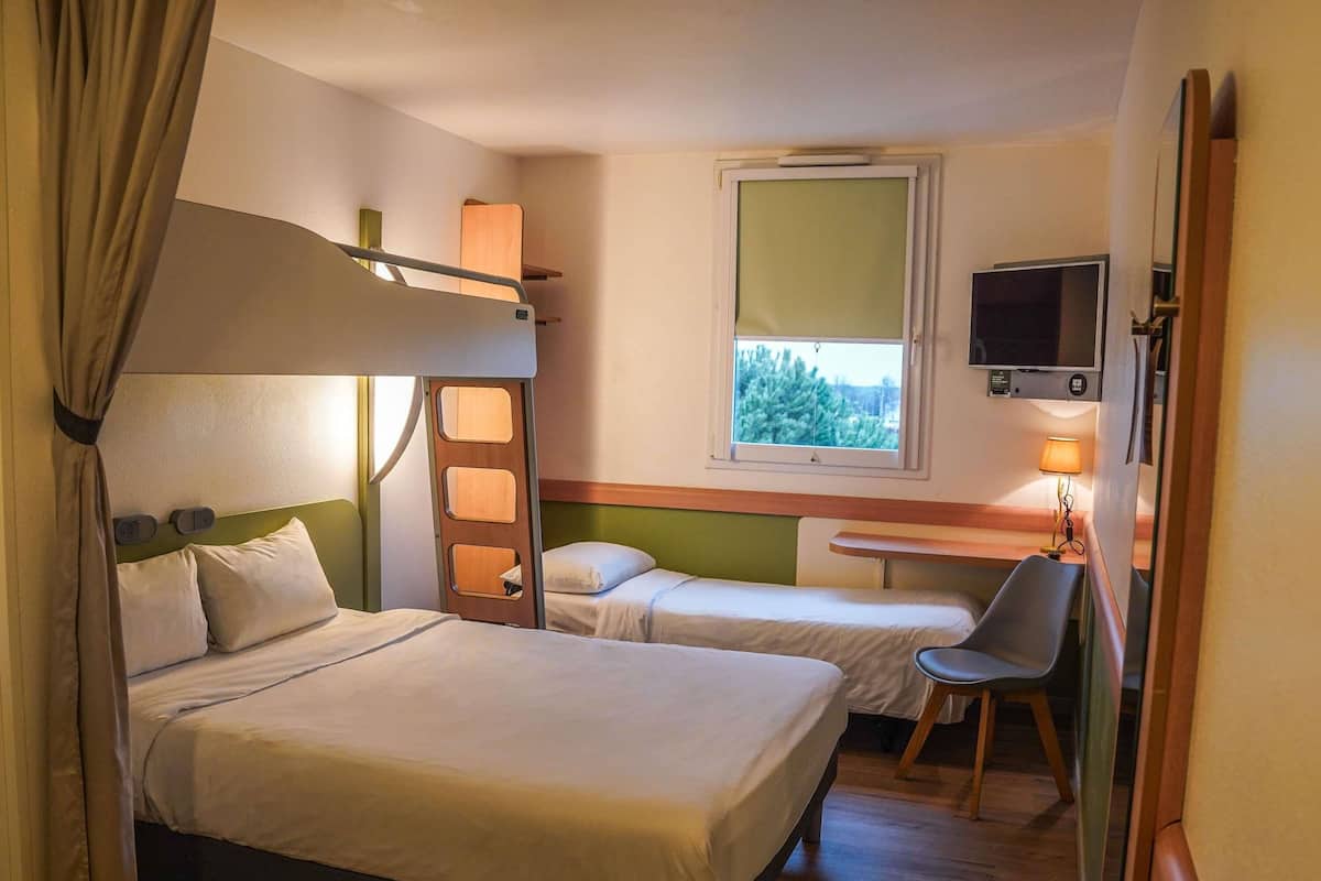 A hotel room with a bunk bed, a single bed, a desk with a chair, a TV, and a window with a view of trees.