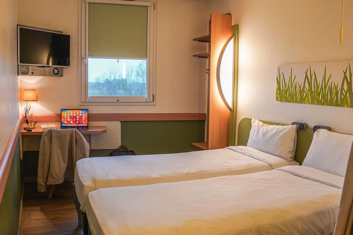 A hotel room with two beds, a desk with a laptop, a TV, and a window with a view of trees.