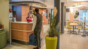 A hotel reception area with a wooden counter, a clock, and a large potted plant. - ibis budget Bayeux (Nonant)