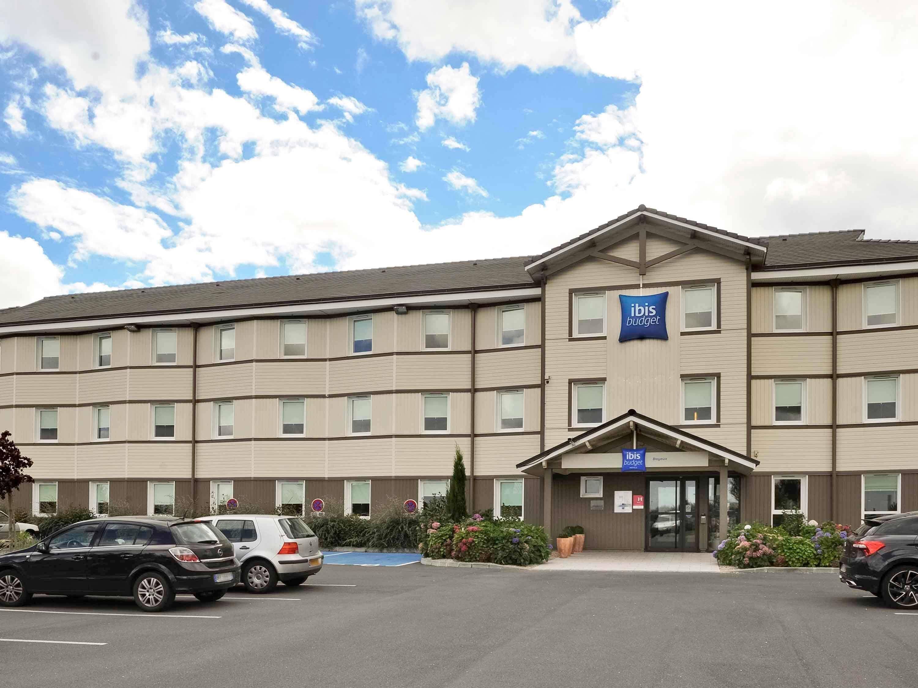 A multi-story hotel with a flat roof, a central entrance, and a parking lot with several cars.