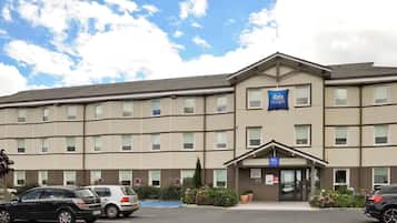 A multi-story hotel with a flat roof, a central entrance, and a parking lot with several cars.