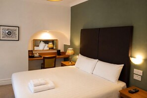 Quarto de hotel com cama, escrivaninha, cadeira, abajur e espelho fixado na parede.