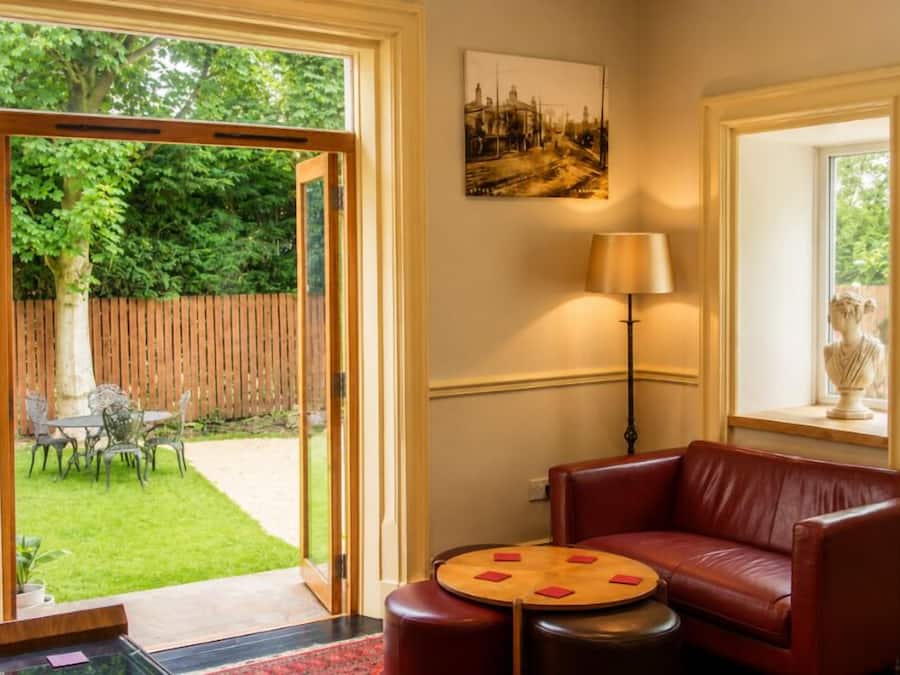 A room with a red leather sofa, a round wooden table, and a lamp. There is a door leading to a garden with a wooden fence and a table outside.