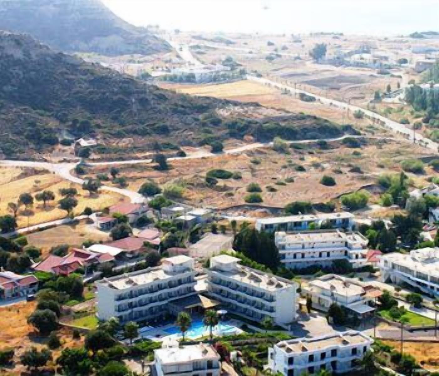 A coastal resort with multiple-story buildings, a central swimming pool, and a mountainous backdrop.