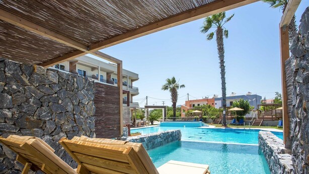 A pool area with wooden lounge chairs, a stone wall, and a clear view of a building in the background. - Dias Studios (Rhodes)