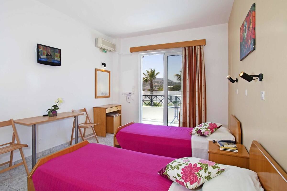 Habitación de hotel con dos camas, un escritorio, una silla y una ventana con vista a palmeras.