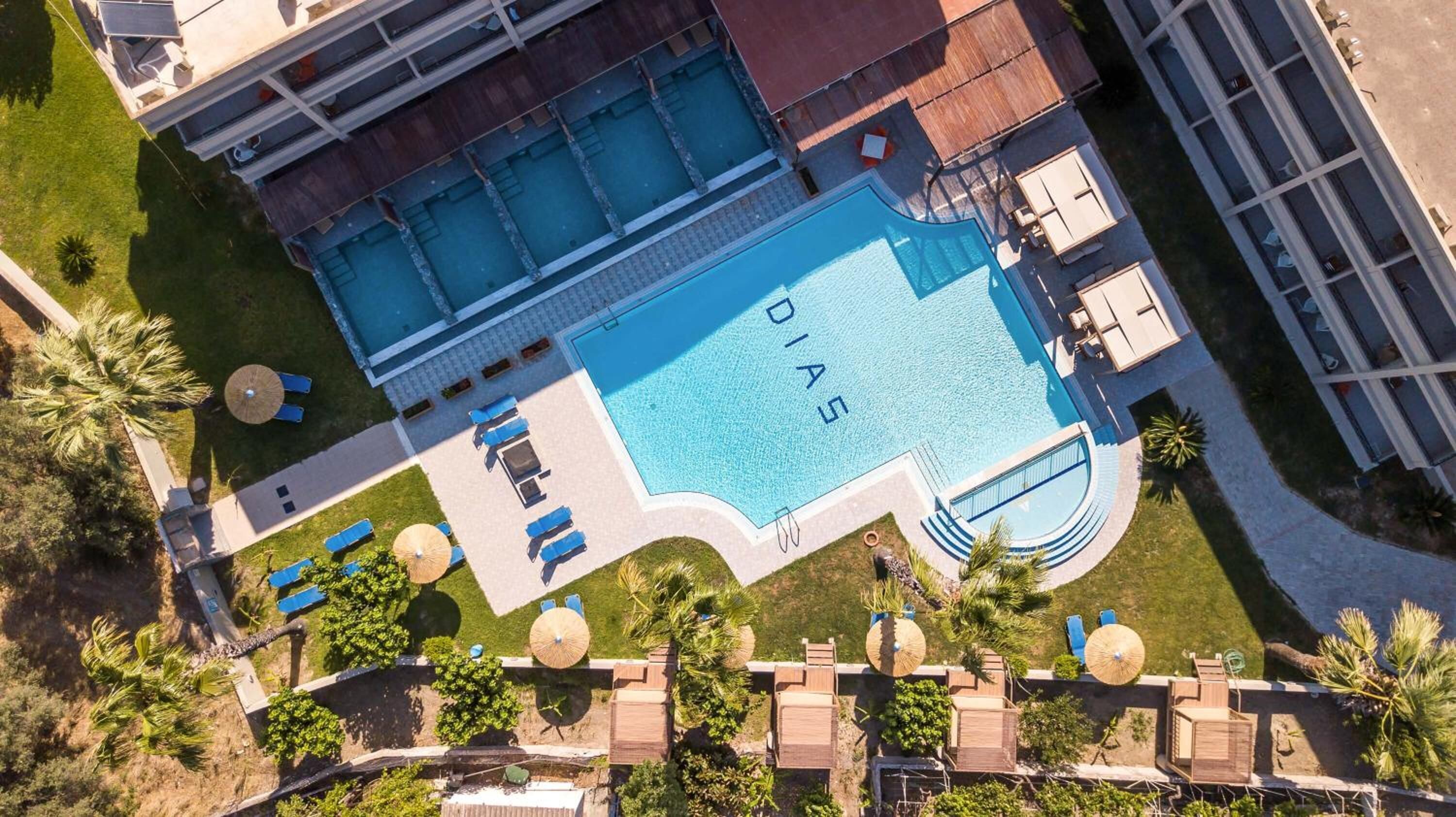 An aerial view of a hotel with a large swimming pool, multiple lounge areas with umbrellas, and a well-maintained garden.