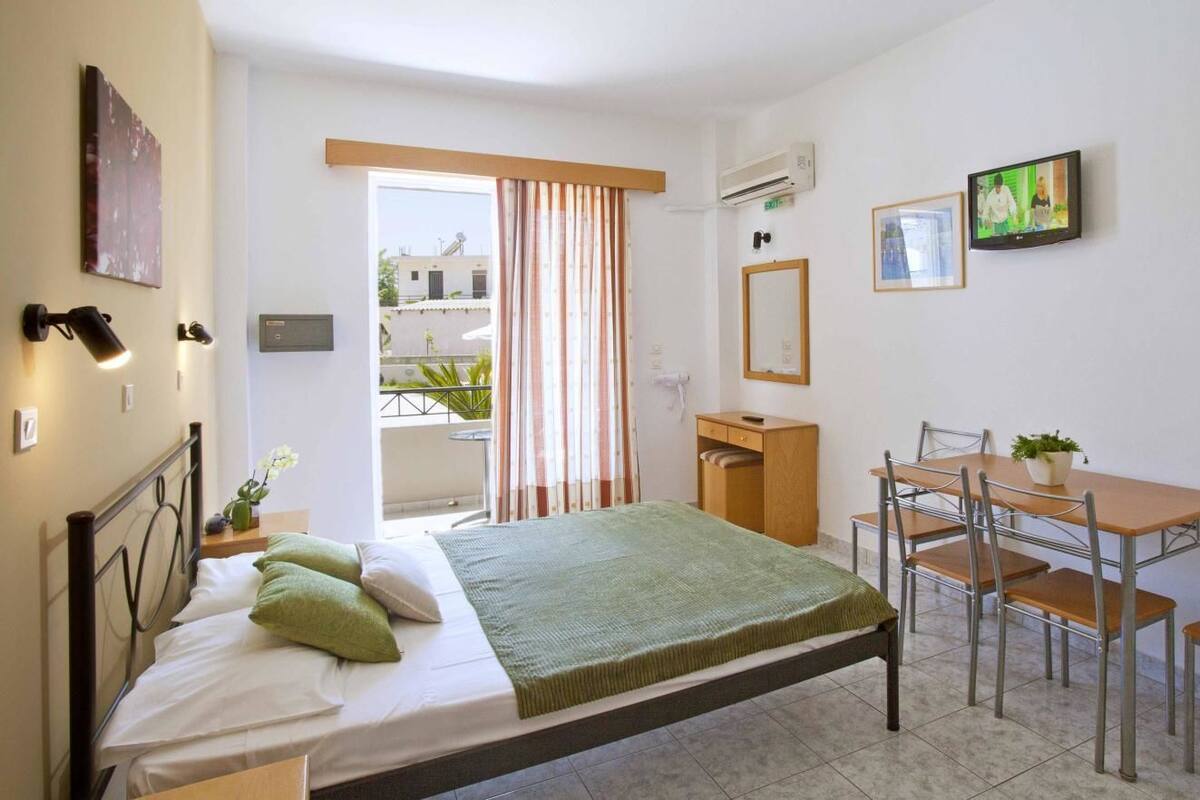 Habitación de hotel con cama, escritorio y silla. Cuenta con una ventana con cortinas, un televisor en la pared y una planta en el escritorio.