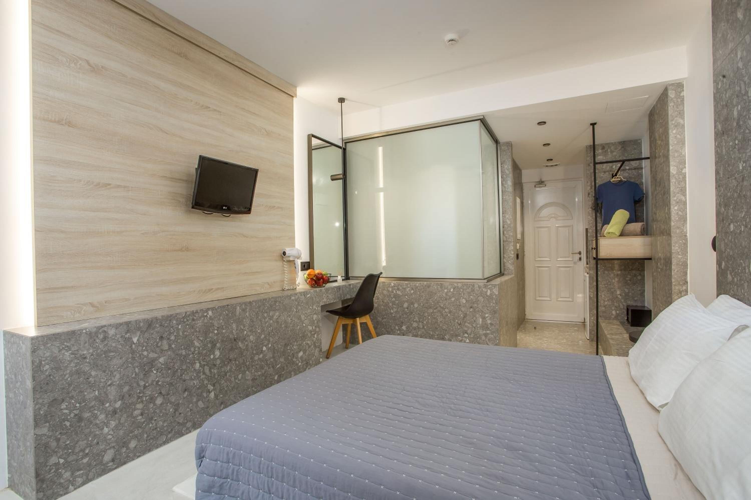 A modern hotel room with a large bed, a desk with a chair, a flat-screen TV mounted on the wall, and a glass shower enclosure.
