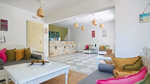 A spacious living room with a white sofa, a white coffee table, and a checkered floor. There is a staircase leading to an upper level, and the room is well-lit with pendant lights. - Camara Hotel (Naxos)