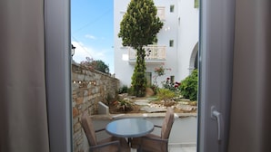 An open door leading to a patio with a round table and two chairs, a stone wall, and a building with balconies and a tree in the background. - Camara Hotel (Naxos)