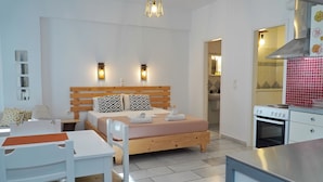 A modern bedroom with a wooden bed, bedside lamps, a white desk, and a kitchenette. - Camara Hotel (Naxos)