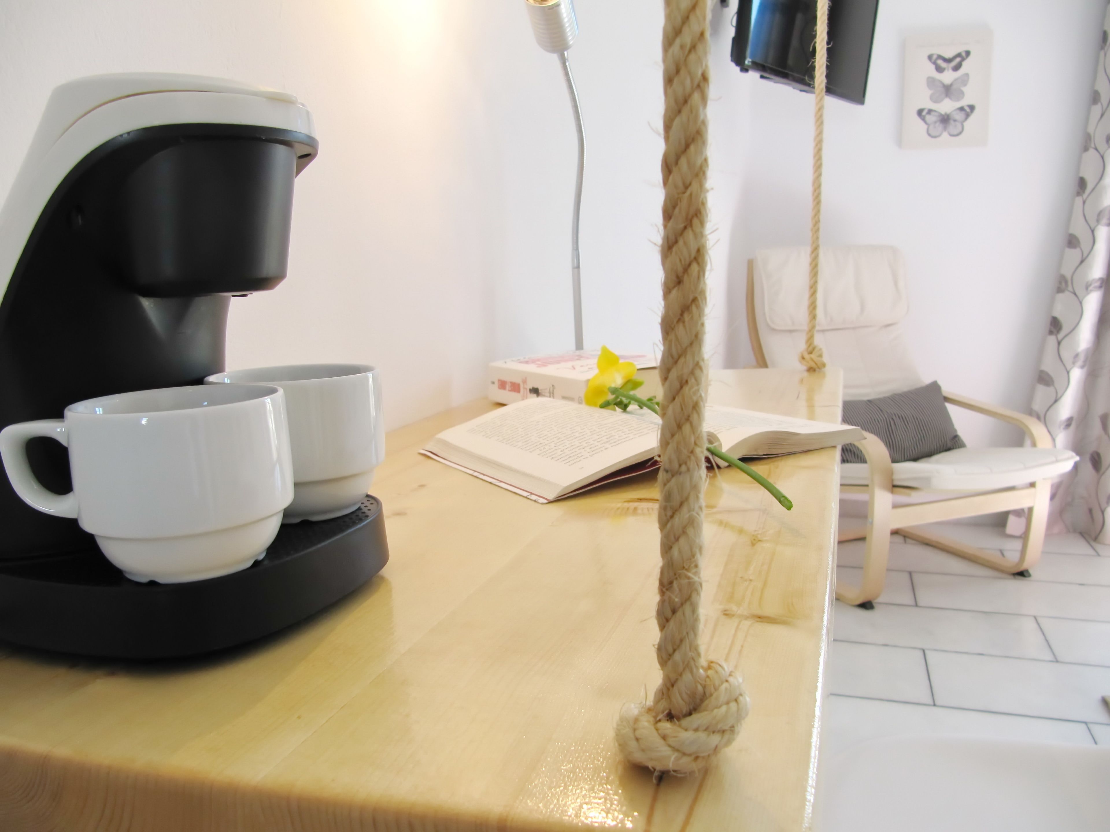 Une machine à café avec deux tasses, un livre, une fleur et une chaise dans une pièce dont le mur est blanc et où se trouve un téléviseur.