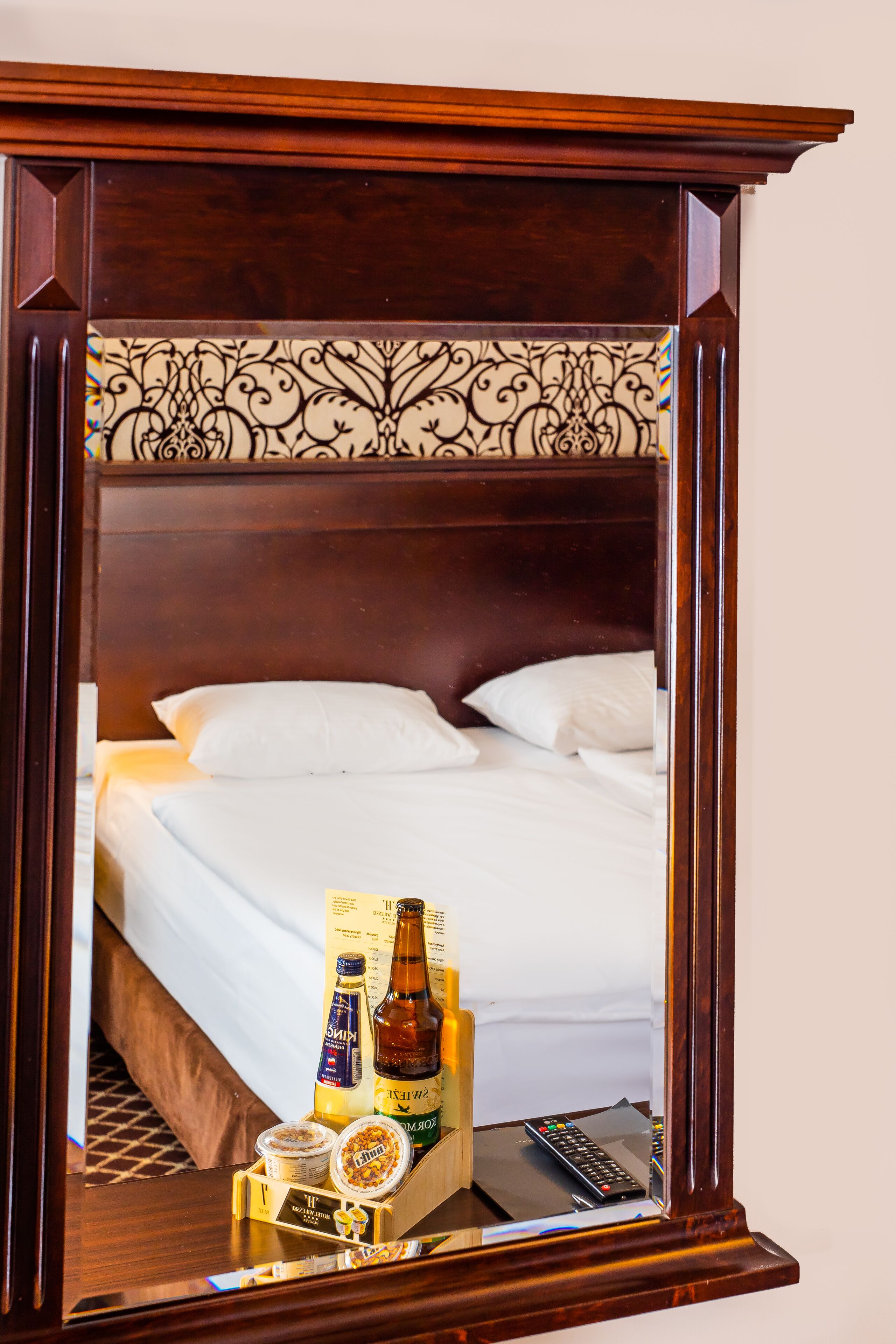 Ein Spiegel spiegelt ein ordentlich bezogenes Bett mit Kissen, einen Nachttisch mit einer Flasche, einer Fernbedienung und einem Tablett mit Snacks wider.