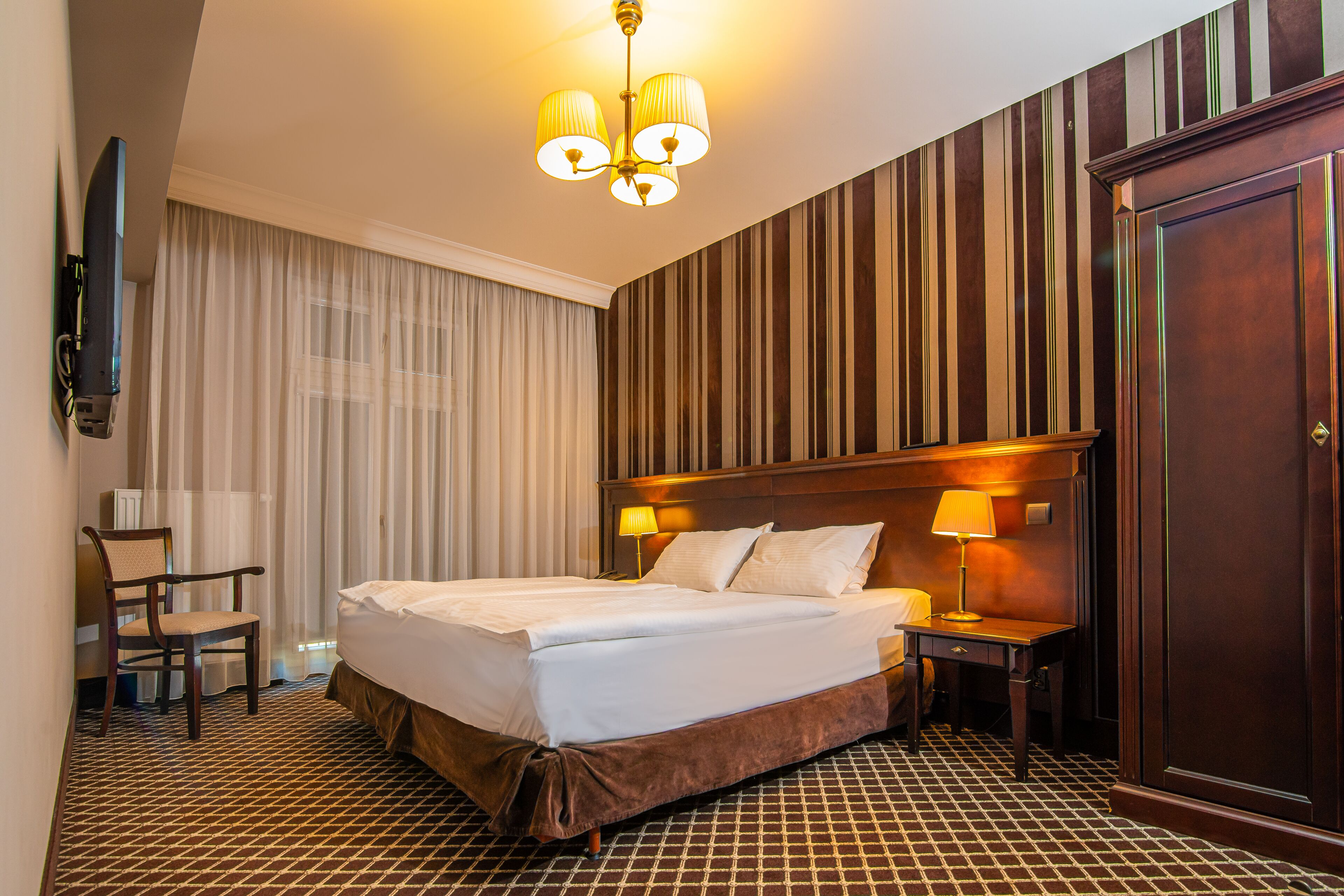 Ein Hotelzimmer mit einem großen Bett, zwei Nachttischen inklusive Lampen, einem Sessel, einem an der Wand montierten Fernseher und einem Holzschrank.