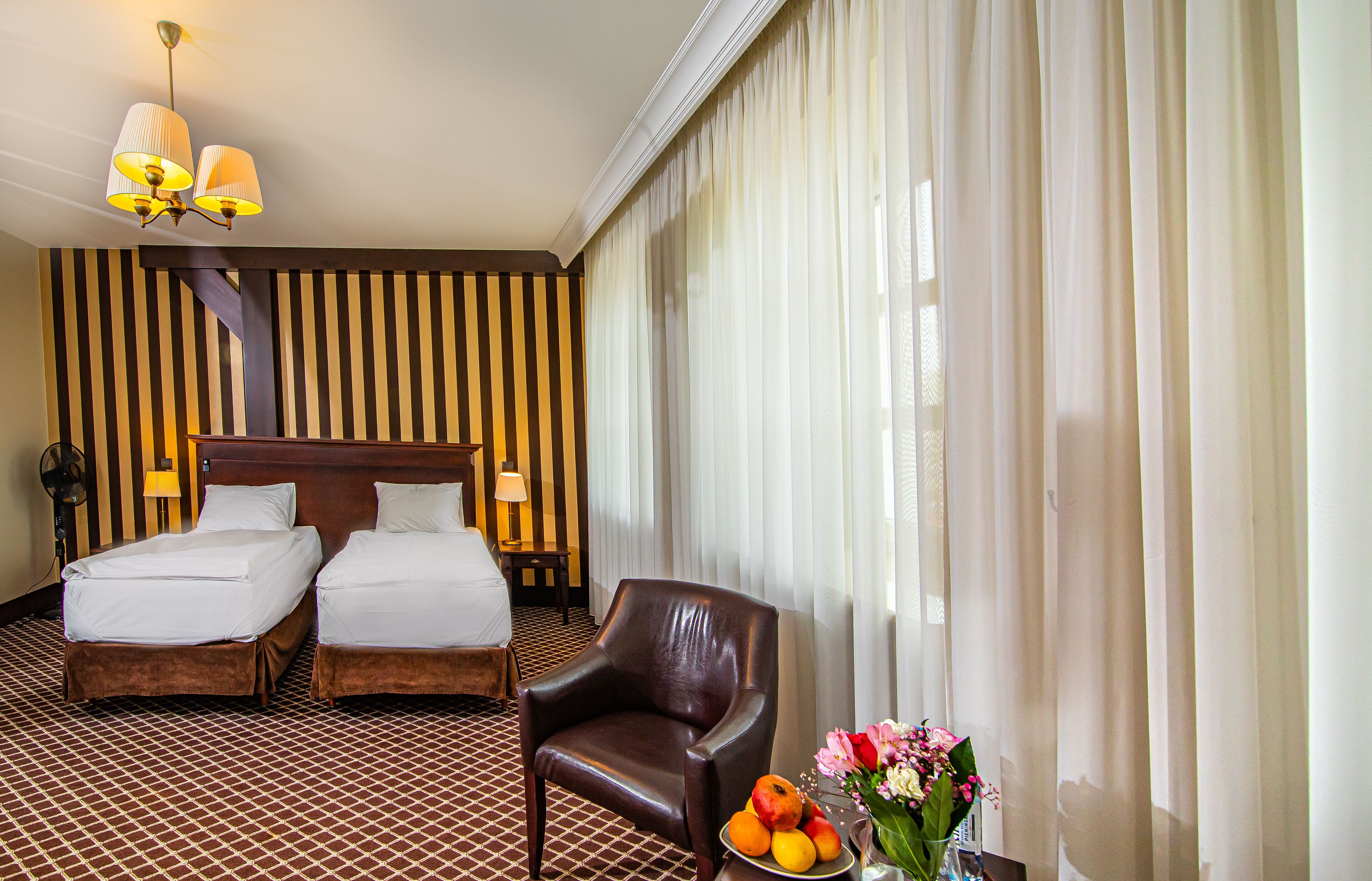 A hotel room with two beds, a wooden headboard, a brown leather chair, a small table with fruit and flowers, and striped wallpaper.