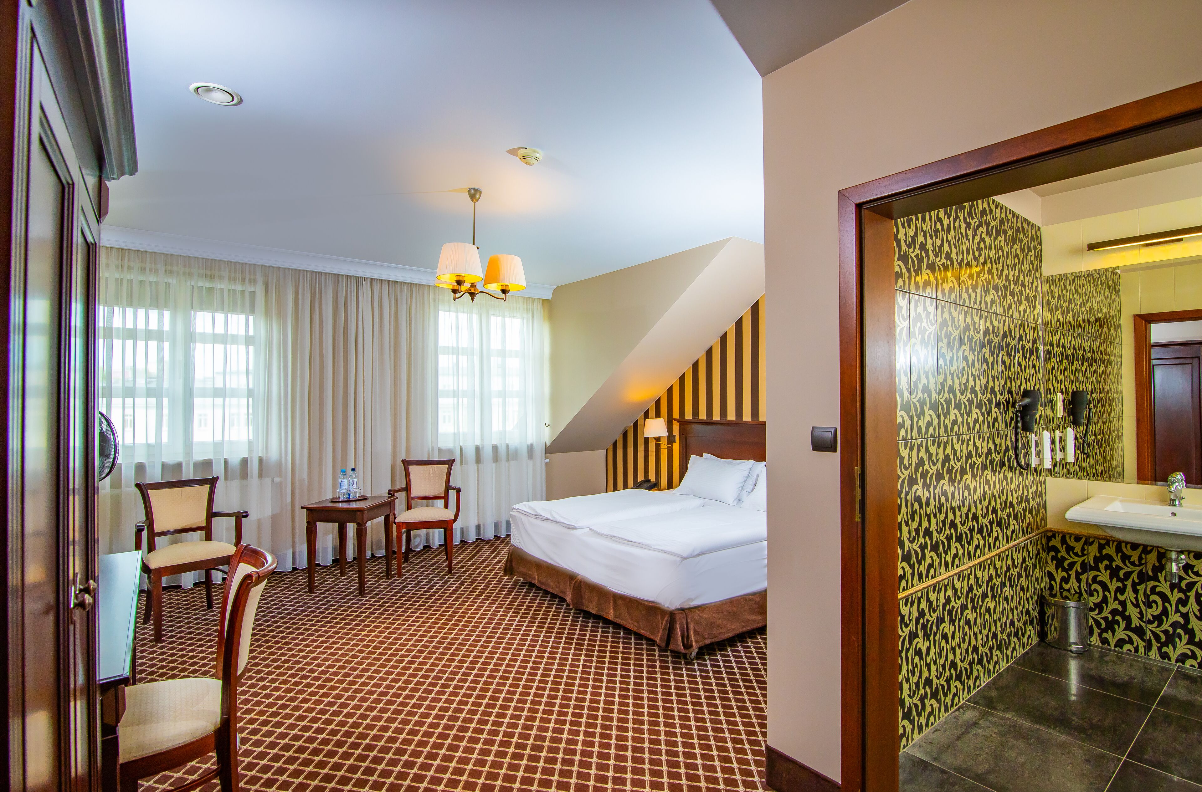 Ein Hotelzimmer mit Bett, Schreibtisch mit Stühlen, Fenster mit Vorhängen und Bad mit Waschbecken und Spiegel.