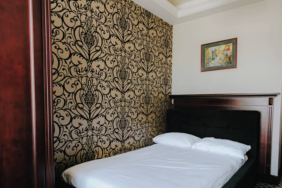 A neatly made bed in a room with patterned wallpaper, a wooden headboard, and a chandelier.
