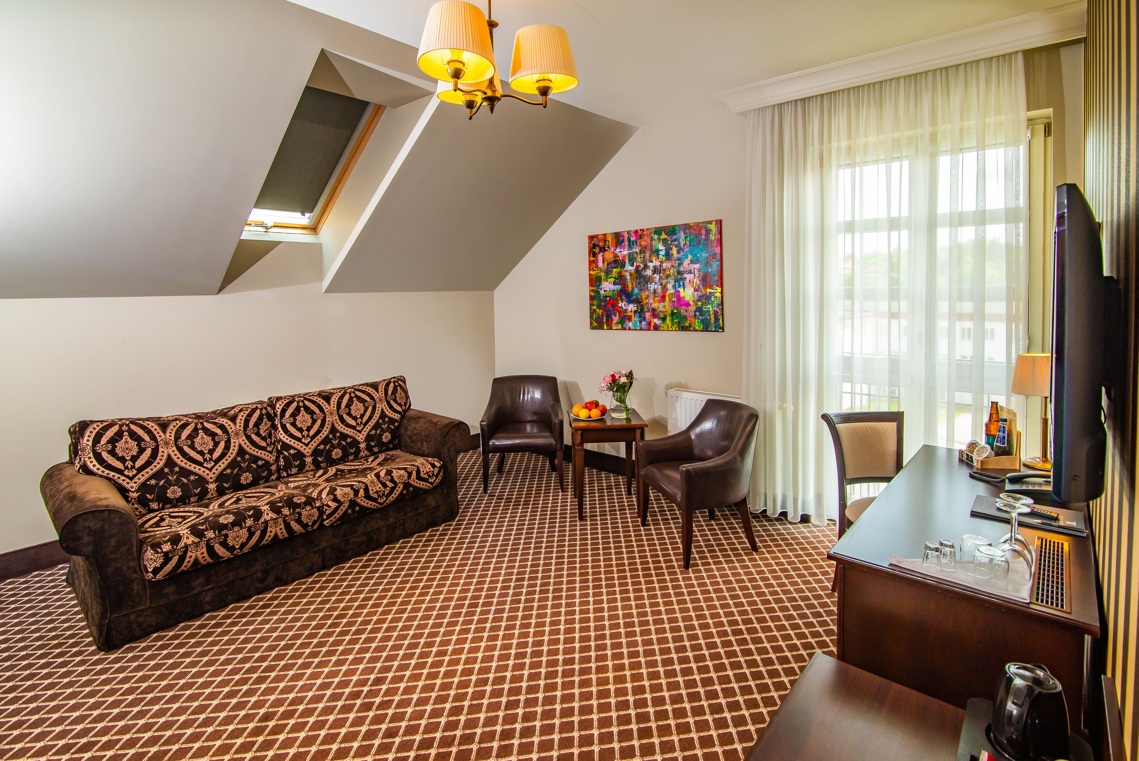 A hotel room with a patterned carpet, a sofa, two chairs, a small table with a vase of flowers, a desk with a computer, and a television.