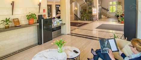 A lobby with a reception desk, two marble tables, and a woman reading a book.