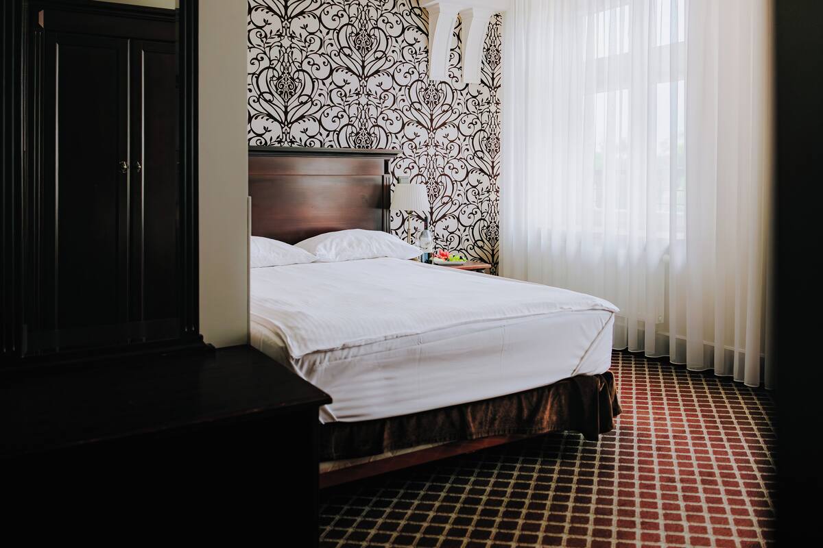 A hotel room with a large bed, a wooden headboard, a nightstand with a lamp, and a patterned wallpaper.