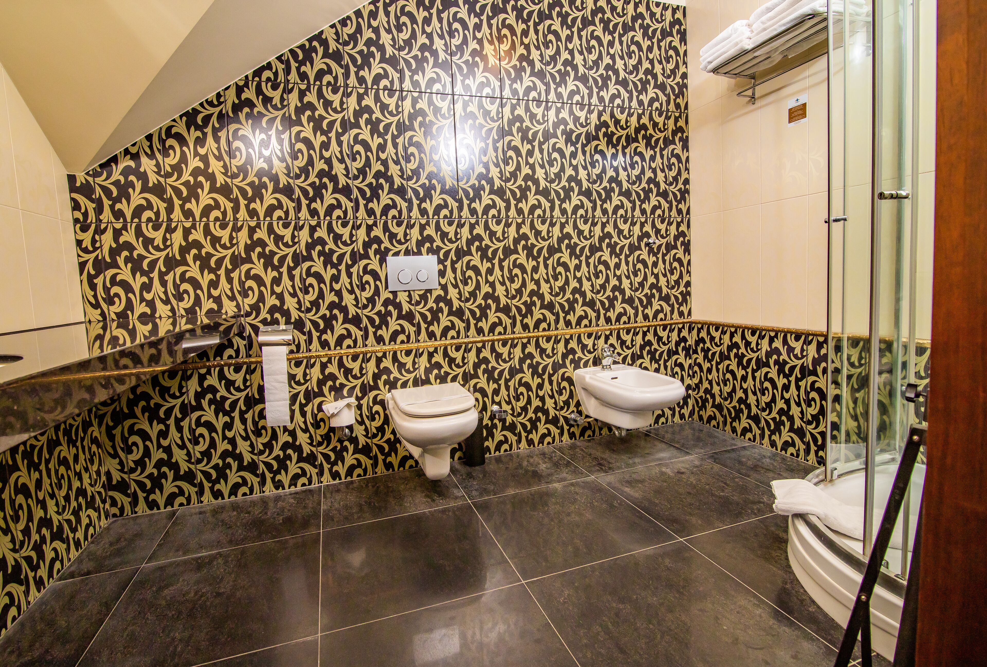 Un baño con paredes de diseño negro y dorado, dos inodoros, un lavabo y una cabina de ducha de vidrio.
