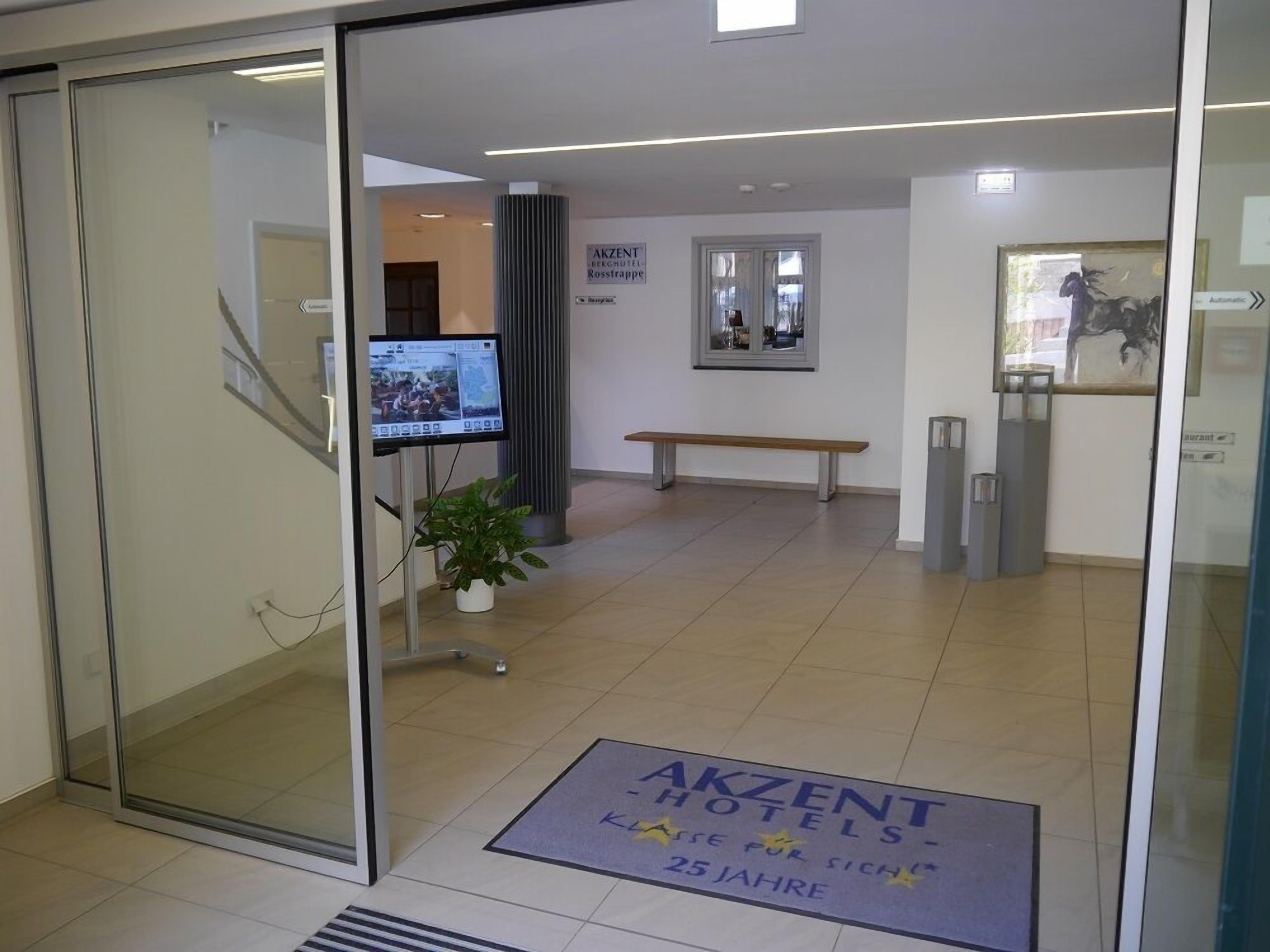 Zona de recepción de un hotel con puerta de vidrio, mostrador de recepción, televisor, banca y felpudo de bienvenida.