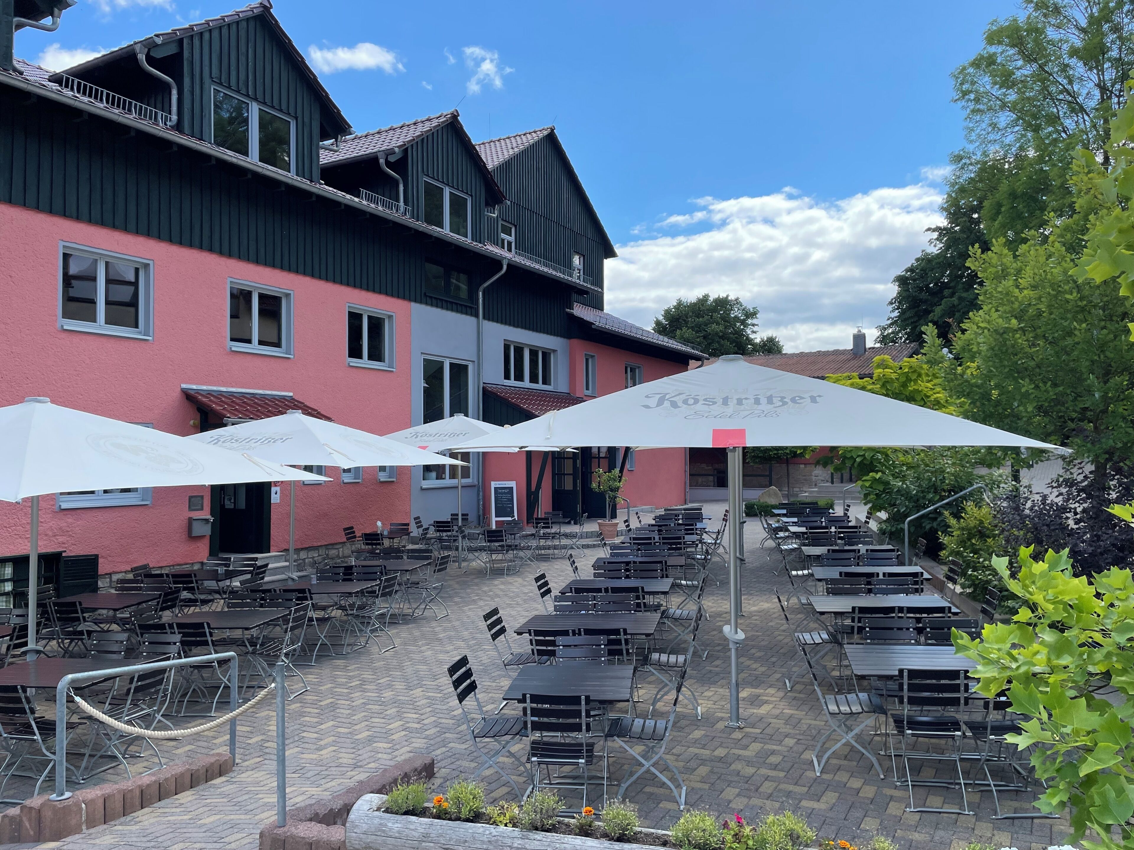 Un espace extérieur aménagé avec des tables et des chaises, des parasols, et un bâtiment à la façade rouge.
