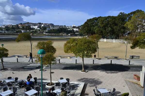 An outdoor cafe with tables and chairs, a paved walkway, and a view of a beach and buildings in the background.