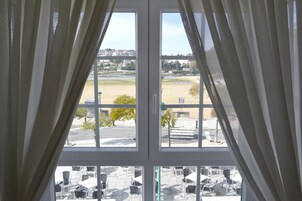 Vista a través de una ventana con cortinas, mostrando un área exterior con mesas y sillas, un campo de césped y edificios al fondo.