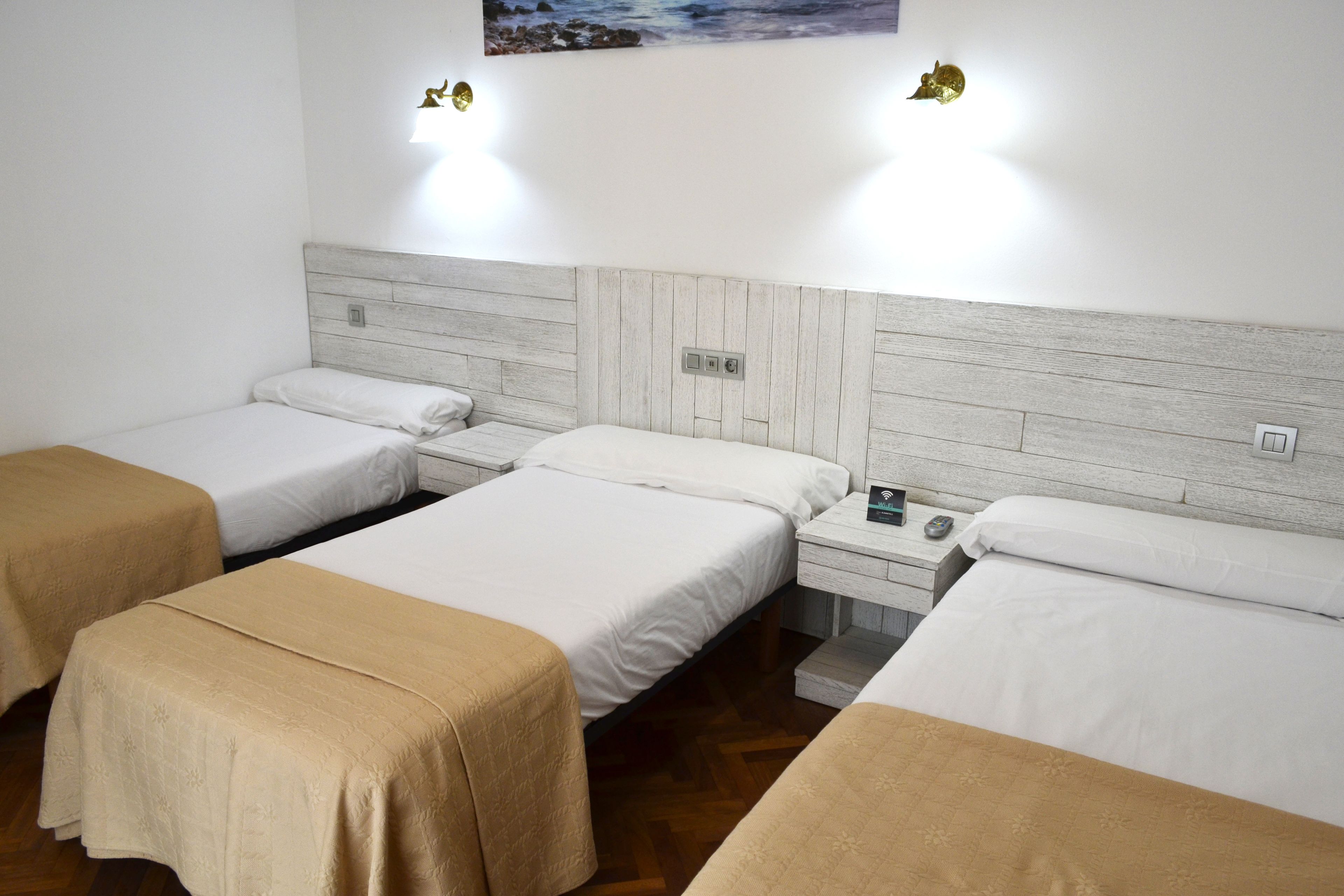 Een hotelkamer met drie eenpersoonsbedden, elk voorzien van een beige sprei en witte lakens. Tussen de bedden staat een nachtkastje met een lamp en een klein tafeltje. De kamer heeft een houten hoofdbord en een schilderij aan de muur.