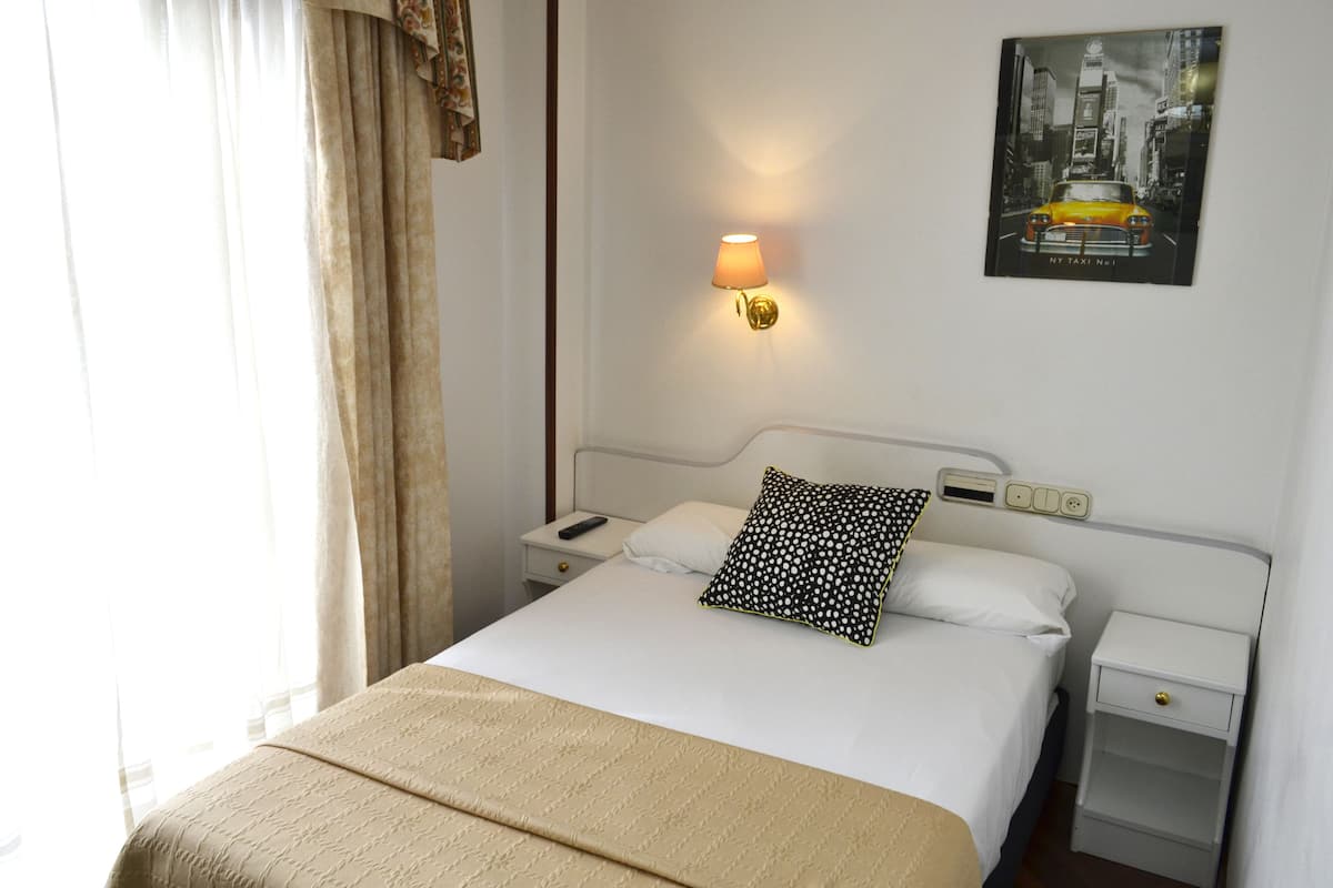 A neatly made bed with a beige bedspread, a black and white patterned pillow, and a white pillow. There is a wall-mounted lamp on the left side of the bed. A framed picture of a yellow taxi is on the wall above the bed.