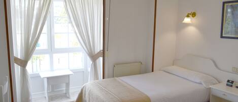 Un dormitorio con cama, una banca, mesita de noche, lámpara y ventana con cortinas.