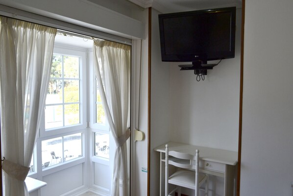 Una habitación con ventana, cortinas, un televisor montado en la pared y una silla blanca.