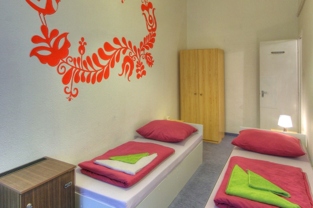 Una habitación con dos camas, un armario de madera, una puerta blanca y una pared decorada con motivos florales rojos.