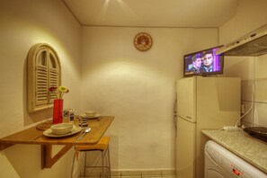 Een compacte keuken met een houten tafel, een kleine koelkast en een televisietoestel dat een programma toont.