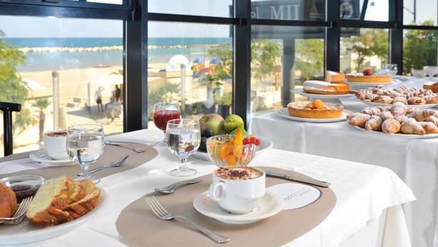 A breakfast buffet with a variety of pastries, fruits, and beverages, set against a backdrop of a beach view through large windows. - Milano Resort (Bellaria-Igea Marina)