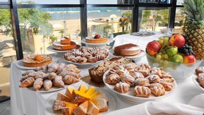 A buffet table with various pastries and fruits, set against a backdrop of a beach view through large windows. - Milano Resort (Bellaria-Igea Marina)
