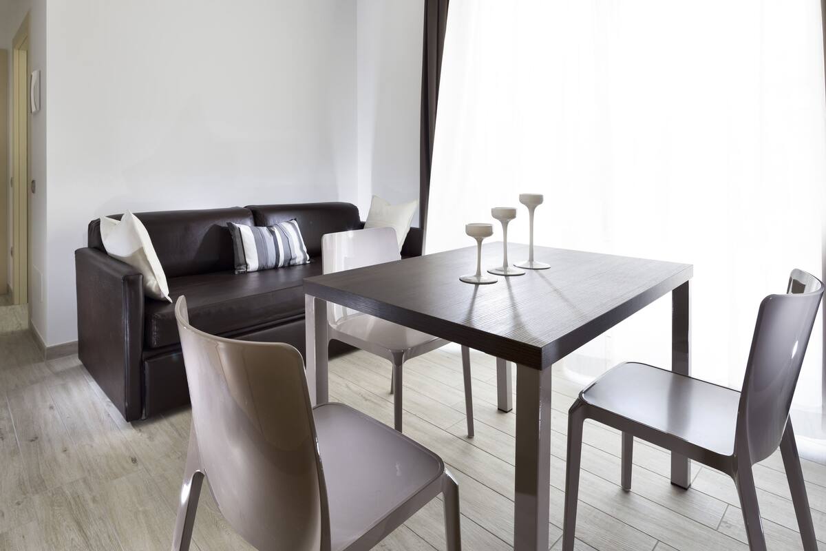 Una sala de estar moderna con un sofá de cuero oscuro, una mesa de comedor de madera con cuatro sillas y un amplio ventanal que proporciona luz natural.